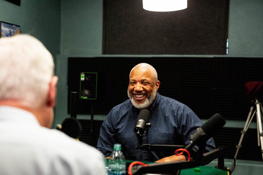 John Register smiles sitting at the podcast interview table with Doug Price